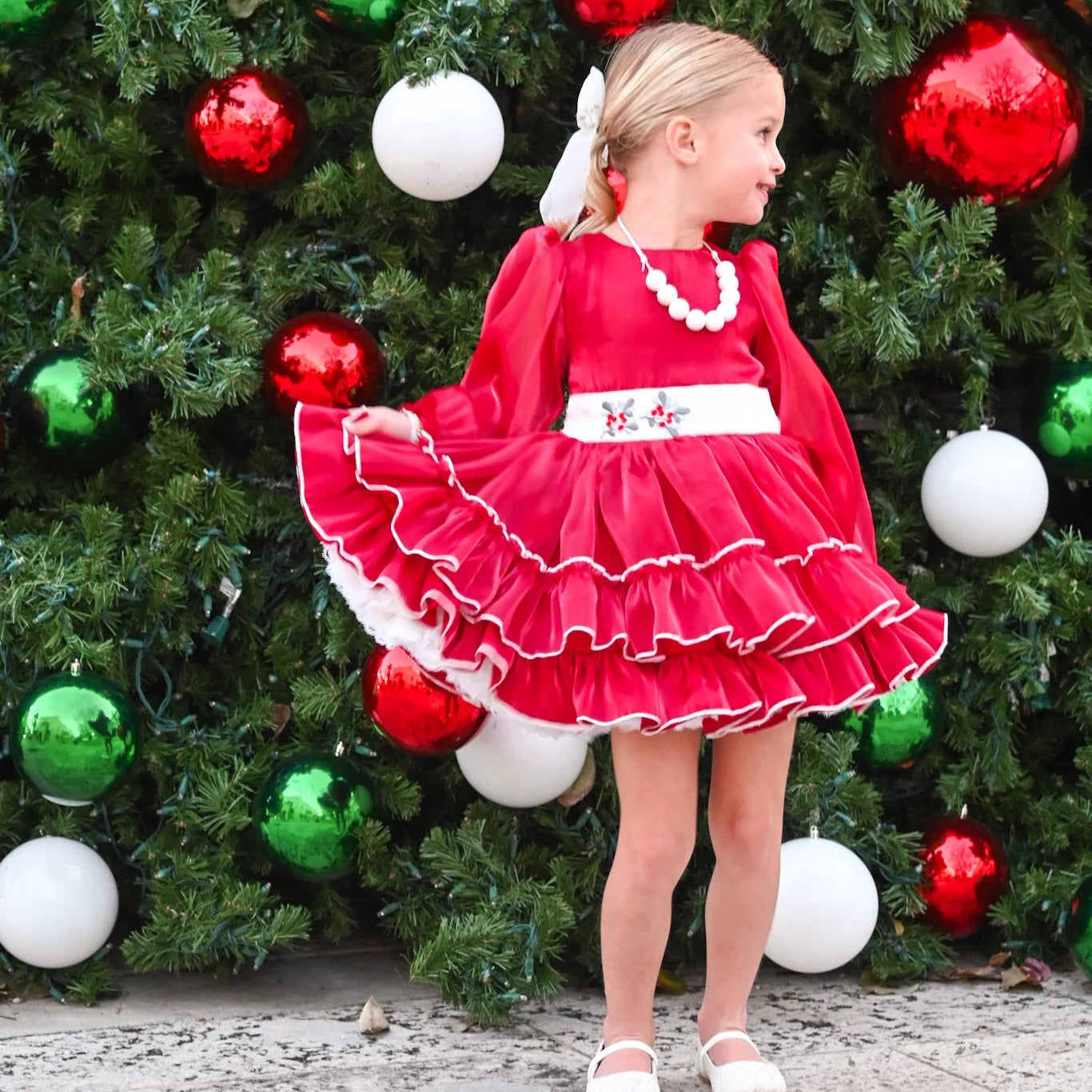 Young girl in a red dress standing in front of a Christmas tree with colorful ornaments.